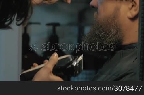 Close up view of female barber taking cares of lush beard of white old man. Barber combing and trimming beard