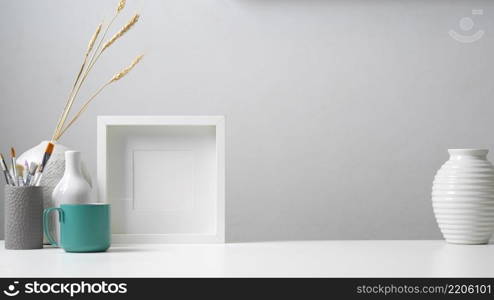 Close up view of home office desk with copy space, mock up frame, paint brushes and decorations in white concept. Home office desk with copy space, mock up frame, paint brushes and decorations in white concept
