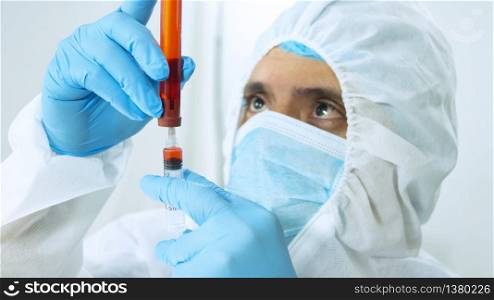 Close up view of latin doctor in bioprotective suit, face mask and blue gloves taking a blood sample in a test tube with a syringe on white background. Close up view of latin doctor in bioprotective suit, mask and blue gloves taking a sample from a test tube with a syringe on white background