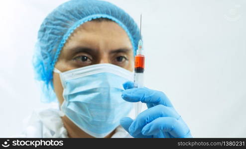 Close up view of latin doctor in bioprotective suit, face mask and blue gloves staring with his eyes at a syringe with a blood sample he is holding with his hand on white background. Latin doctor in bioprotective suit, face mask and blue gloves staring with his eyes at a syringe with a blood sample he is holding with his hand on white background