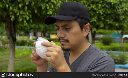 Close up view of latin man on the street staring at a white face mask he is holding with both hands about to put it on. Latin man on the street staring at a white face mask he is holding with both hands about to put it on