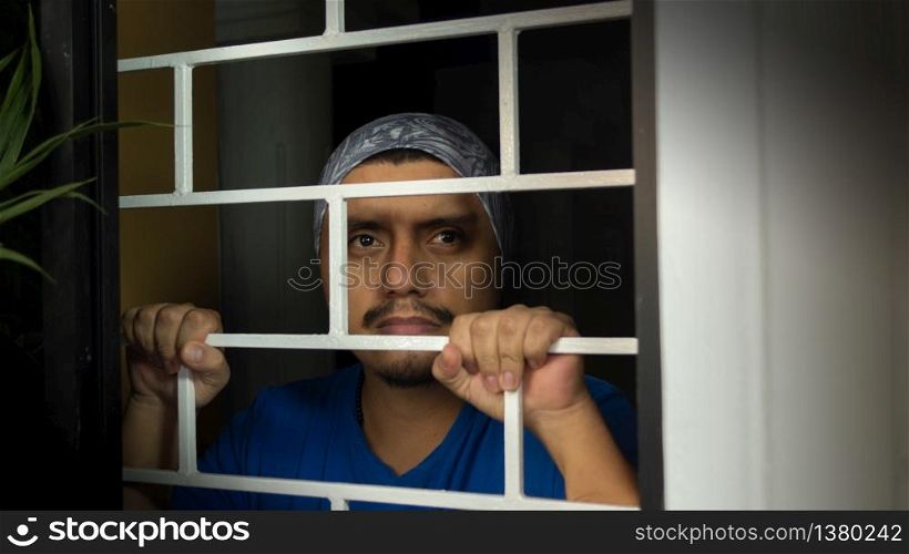 Close up view of latin man with blue shirt locked in his house looking out through the window, leaning against security bars. Latin man with blue shirt locked in his house looking out through the window, leaning against security bars