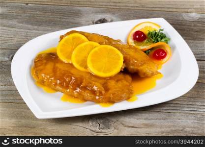 Close up view of lemon chicken with tangy sauce and slice lemons on top in white plate with rustic wood underneath.