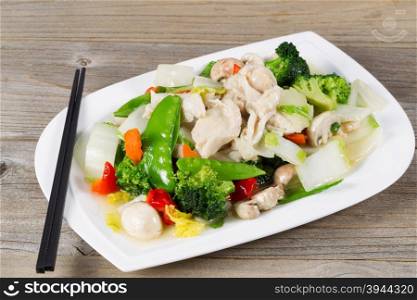 Close up view of stir fried white chicken pieces with broccoli, snow peas, peppers and mushroom in white plate on rustic wood setting.