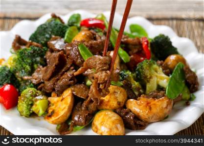 Close up view of tender beef slices, mushrooms, broccoli, peppers and peas in white plate. Selective focus on single piece held by chopsticks.