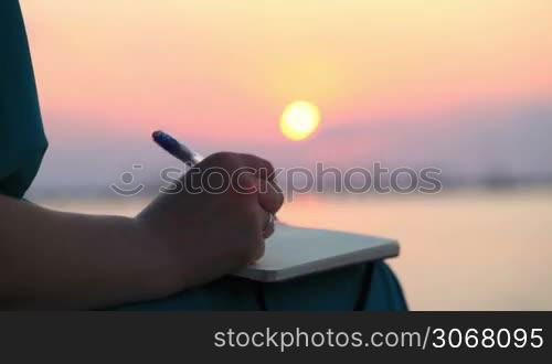 Close up view of the hand of a woman writing in her diary at sunset with the glowing orb of the sun reflected over a still ocean