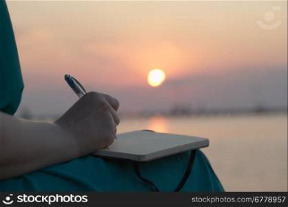 Close up view of the hand of a woman writing in her diary at sunset with the glowing orb of the sun reflected over a still ocean