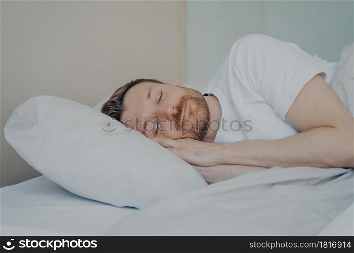 Close up view photo of bearded young male with eyes closed fall asleep on his vacation on sunday, sleeping with smile while lying on white pillow in bedroom. Sweet dreaming and rest concept. Close up view photo of bearded young male fall asleep in his bed