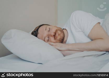 Close up view photo of bearded young male with eyes closed fall asleep on his vacation on sunday, sleeping with smile while lying on white pillow in bedroom. Sweet dreaming and rest concept. Close up view photo of bearded young male fall asleep in his bed