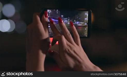 Close up woman&acute;s hand holding a smartphone and taking photo of night city on the street. Shallow depth of field. Focus on image in phone. Background cityscape and streetlights.