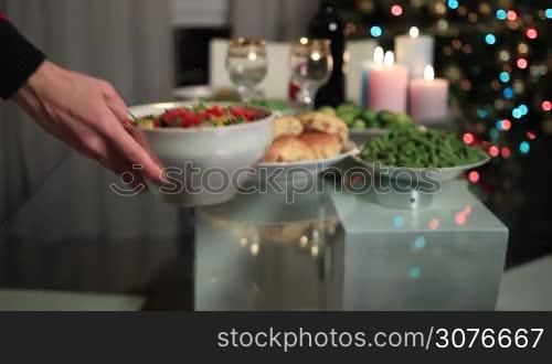 Close up woman&acute;s hand serving bowl of salad on festive table over blurred decorated Christmas table setting and Christmas tree bakground. Family celebrating Christmas Eve together at home.