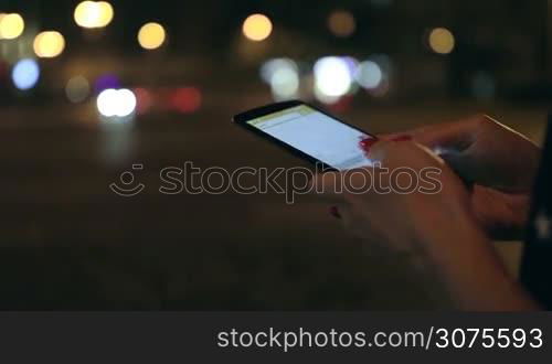 Close up woman&acute;s hands texting on mobile phone on the street at night. Background night city.