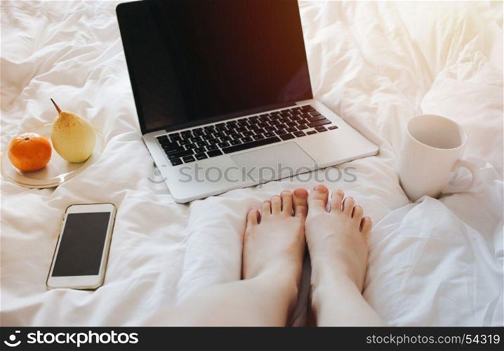 Close up Woman feet relaxing on bed with a laptop computer, smartphone, tea and fruits on white bed sheet