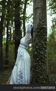 Close up woman in embroidered dress leaning against tree trunk concept photo. Side view photography with forest on background. High quality picture for wallpaper, travel blog, magazine, article. Close up woman in embroidered dress leaning against tree trunk concept photo