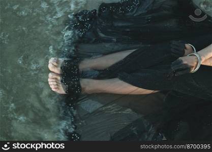 Close up woman legs concept photo. Beautiful black dress. Side view fine art photography with sea tide on background. High quality picture for wallpaper, travel blog, magazine, article. Close up woman legs concept photo