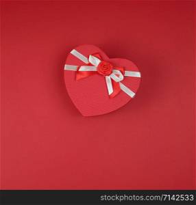 closed red gift box in the form of a heart with a bow on a red background, copy space, flat lay
