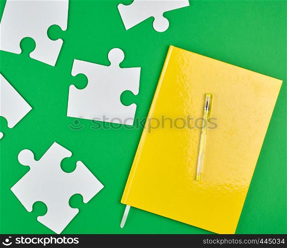 closed yellow notebook and pen, next to it are large empty white paper puzzles, green background, top view