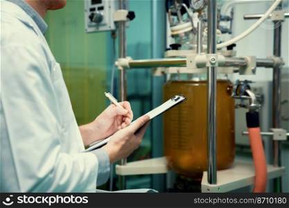 Closeup apothecary scientist using a clipboard and pen to record information from a CBD oil extractor and a scientific machine used to create medicinal cannabis products.. Closeup apothecary scientist recording data from CBD oil extractor in laboratory