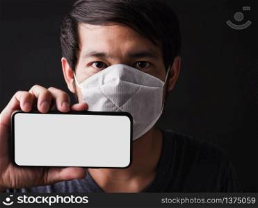Closeup Asian handsome man wearing protective face mask fear in eye holding show mobile phone blank screen near face, hygiene prevention COVID-19 or coronavirus protection concept, black background