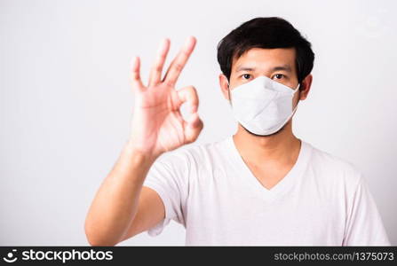 Closeup Asian handsome Man wearing surgical hygienic protective cloth face mask against coronavirus and show OK sign, studio shot isolated white background, COVID-19 medical concept