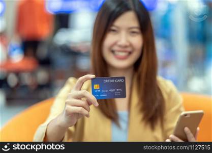 Closeup Asian woman hand holding the credit card and presenting the mobile phone for online shopping over the clothes shop store background, technology money wallet and online payment concept