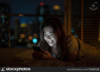 Closeup Asian woman sleeping and using smart mobile phone for social network or video conference call and face time to her lovers on bed over blurred city at night time, lifestyle and relax concept