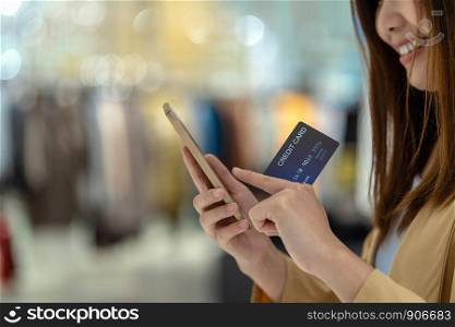 Closeup Asian woman using credit card with mobile for online shopping in department store over the clothes shop store background, technology money wallet and online payment concept, credit card mockup