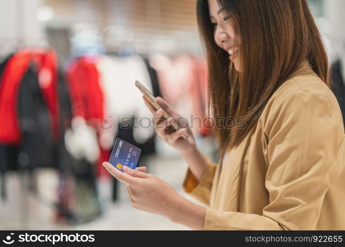 Closeup Asian woman using credit card with mobile for online shopping in department store over the clothes shop store background, technology money wallet and online payment concept, credit card mockup