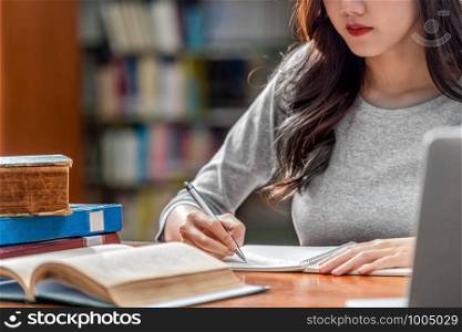 Closeup Asian young Student hand writing homework and using technology laptop in library of university or colleage with various book and stationary over the book shelf background, Back to school