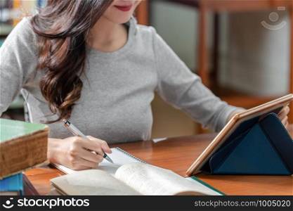 Closeup Asian young Student hand writing homework and using technology tablet in library of university or colleage with various book and stationary over the book shelf background, Back to school