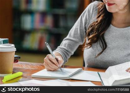 Closeup Asian young Student hand writing homework in library of university or colleage with various book and stationary with coffee cup on wooden table over the book shelf background,Back to school