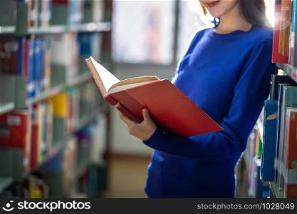 Closeup Asian young Student in casual suit standing and hand holding the book and reading at book shelf in library of university or colleage with various book background, Back to school concept
