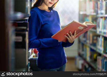 Closeup Asian young Student in casual suit standing and hand holding the book and reading at book shelf in library of university or colleage with various book background, Back to school concept