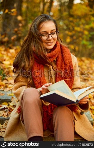 Closeup autumn portrait of young smart woman in eyeglasses and warm clothes reading book outdoors. Closeup of young woman reading book outdoors