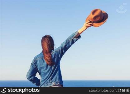 Closeup back view of woman in jeans jacket holding hat and standing and looking at blue ocean and sky.