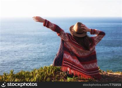 Closeup back view of woman in travel clothes and hat sitting and looking at blue ocean and sky. Travel concept photo. Closeup back view of woman in travel clothes and hat sitting and looking at blue ocean and sky.