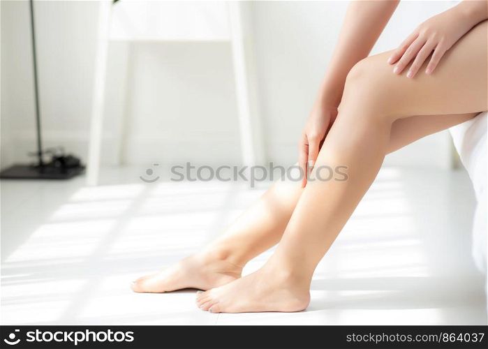 Closeup beautiful young asian woman sitting on a bed stroking legs with soft smooth skin in the bedroom, girl applying body cream and lotion with treatment care, healthy and wellness concept.