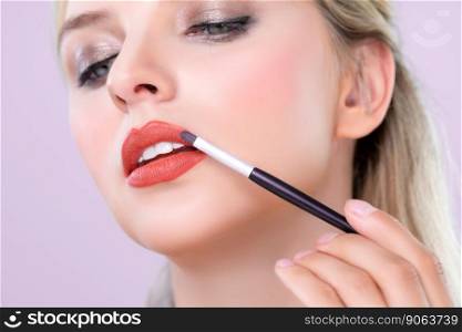 Closeup beautiful young woman with flawless healthy skin and natural makeup putting alluring fashion glossy red lipstick on her lip with lip brush in pink isolated background.. Closeup beautiful young woman putting alluring fashion glossy red lipstick.