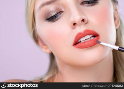 Closeup beautiful young woman with flawless healthy skin and natural makeup putting alluring fashion glossy red lipstick on her lip with lip brush in pink isolated background.. Closeup beautiful young woman putting alluring fashion glossy red lipstick.