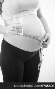Closeup black and white photo of pregnant woman holding glass of water and measuring big belly