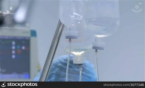 Closeup bottle of liquid drug with lines and needle inside called drop counter in surgery room with blurred heart rate monitor in background. Medical equipment and tools in operating theater.