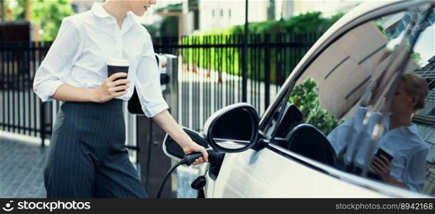 Closeup businesswoman holding coffee, insert EV charger to electric vehicle at public charging station. Eco-friendly car using clean and renewable energy for progressive lifestyle in city.. Closeup progressive businesswoman with coffee recharge EV car.