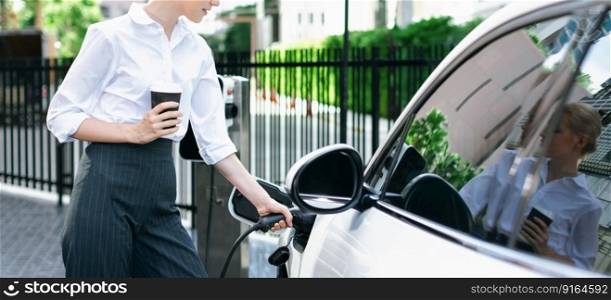 Closeup businesswoman holding coffee, insert EV charger to electric vehicle at public charging station. Eco-friendly car using clean and renewable energy for progressive lifestyle in city.. Closeup progressive businesswoman with coffee recharge EV car.