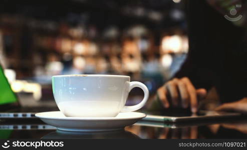 Closeup coffee cups over the photo blurred of asian woman using the smart mobile phone with happiness action in the coffee shop or book store which have low light, drink and backgroup concept