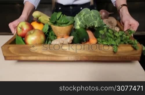 Closeup colorful set of different fresh fruits and raw organic vegetables in wooden tray on the kitchen table. Female hands offering tray with food ingredients for preparing vegan smoothie. Healthy vegetarian diet for weight loss and detox