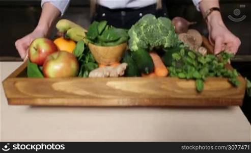 Closeup colorful set of different fresh fruits and raw organic vegetables in wooden tray on the kitchen table. Female hands offering tray with food ingredients for preparing vegan smoothie. Healthy vegetarian diet for weight loss and detox
