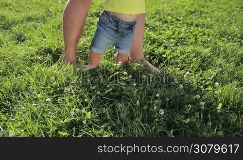 Closeup dad&acute;s and infant child&acute;s feet walking barefoot on green grass in the park on sunny summer day. Father teaching his baby boy to walk on park lawn. Low view. First steps of a child. Slow motion. Steadicam stabilized shot.