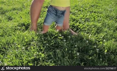 Closeup dad&acute;s and infant child&acute;s feet walking barefoot on green grass in the park on sunny summer day. Father teaching his baby boy to walk on park lawn. Low view. First steps of a child. Slow motion. Steadicam stabilized shot.