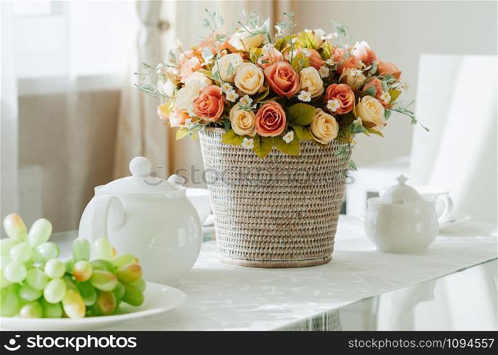closeup dining table setup with white porcelain crockery, cups and pot served for tea, grapes, basket with flowers, natural light from window. Modern comfy interior, show room, restaurant, cafe