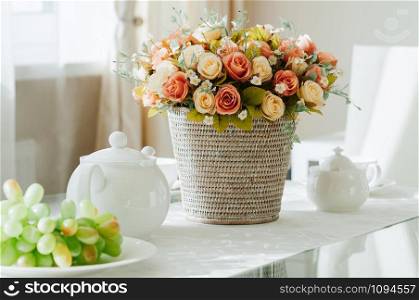 closeup dining table setup with white porcelain crockery, cups and pot served for tea, grapes, basket with flowers, natural light from window. Modern comfy interior, show room, restaurant, cafe